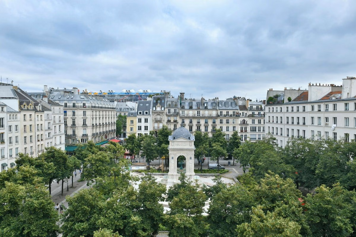 Tree-lined Paris square with a stone fountain and surrounding buildings under a cloudy sky.
